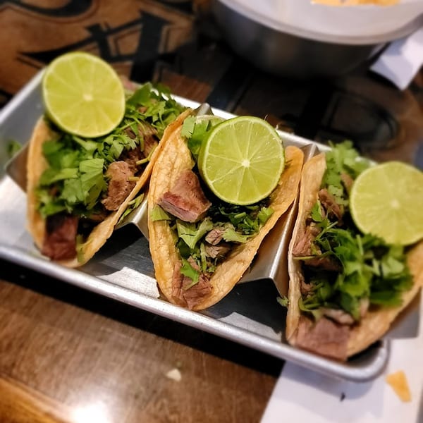 Three carne asada street tacos with cilantro and lime wedges on a metal tray.