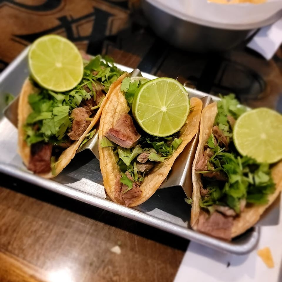 Three carne asada street tacos with cilantro and lime wedges on a metal tray.