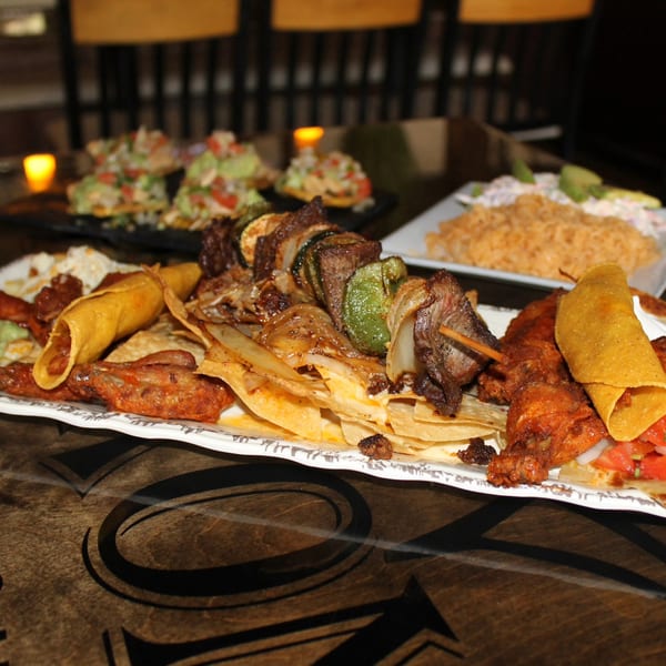 A large platter of Mexican food, including tacos, skewers, and nachos, on a dark wooden table.