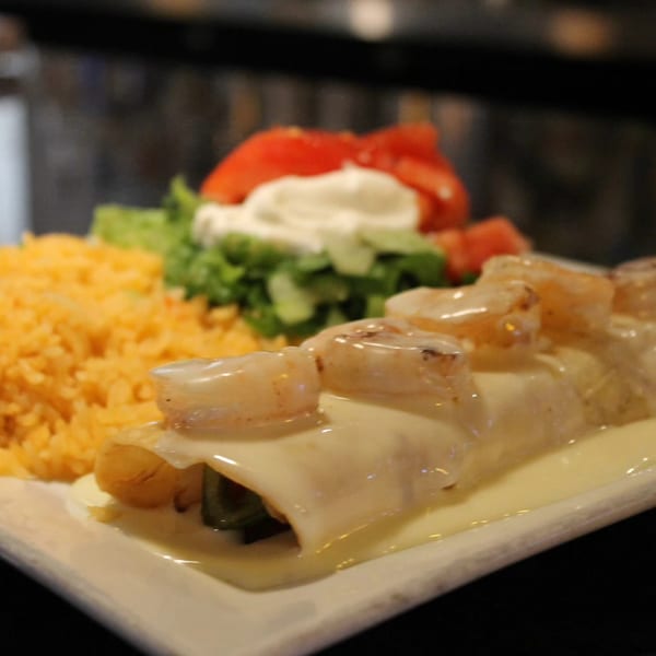 Shrimp enchiladas with white sauce, rice, lettuce, tomato, and sour cream on a white plate.