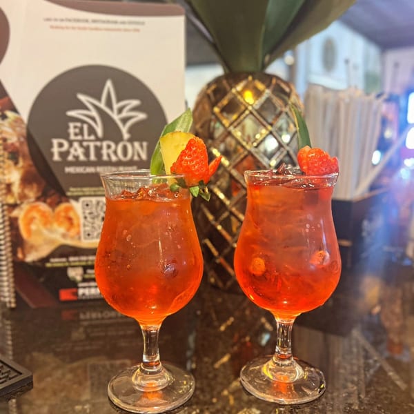 Two red cocktails garnished with strawberries and pineapple, with an El Patron menu in the background.