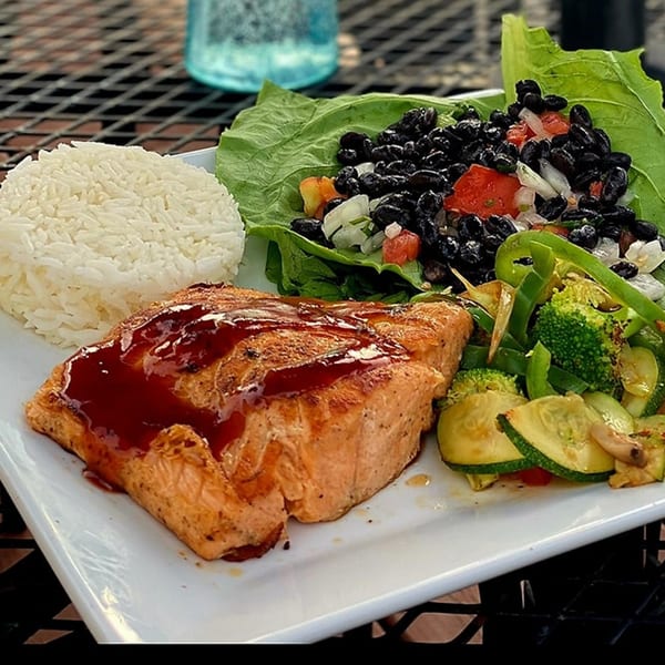 A salmon fillet with sauce, rice, black bean salad, and mixed vegetables on a white plate.