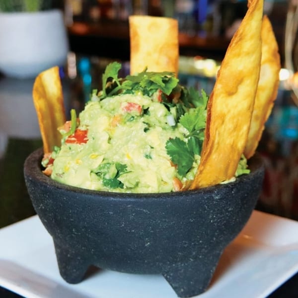 Fresh guacamole with cilantro and diced tomatoes in a dark stone bowl with tortilla chips.